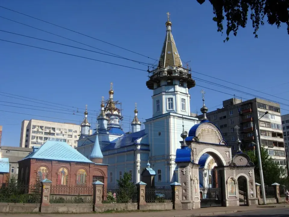 Кирпичный храм с голубыми куполами и шатровой колокольней среди зелени